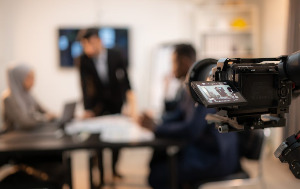 Camera rig filming a meeting in a conference room, with people blurred in the background and a monitor visible on the camera
