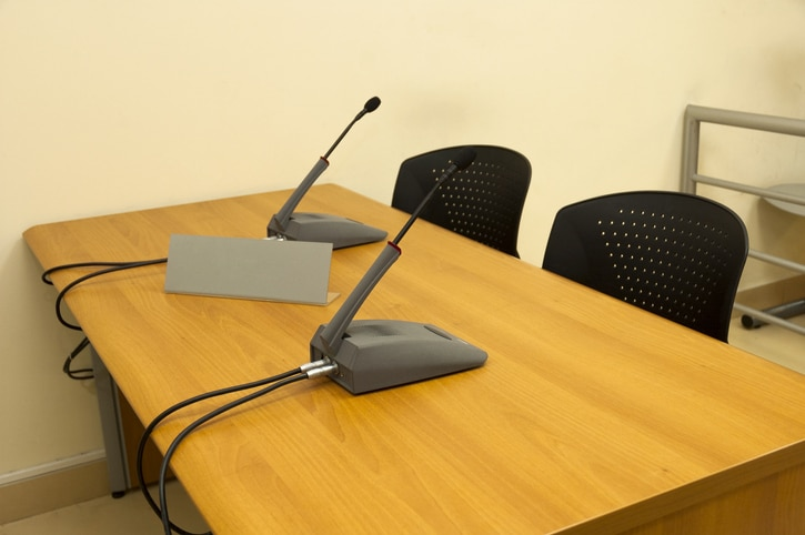 Two gooseneck conference microphones on a wooden table in a meeting room with black perforated chairs nearby.